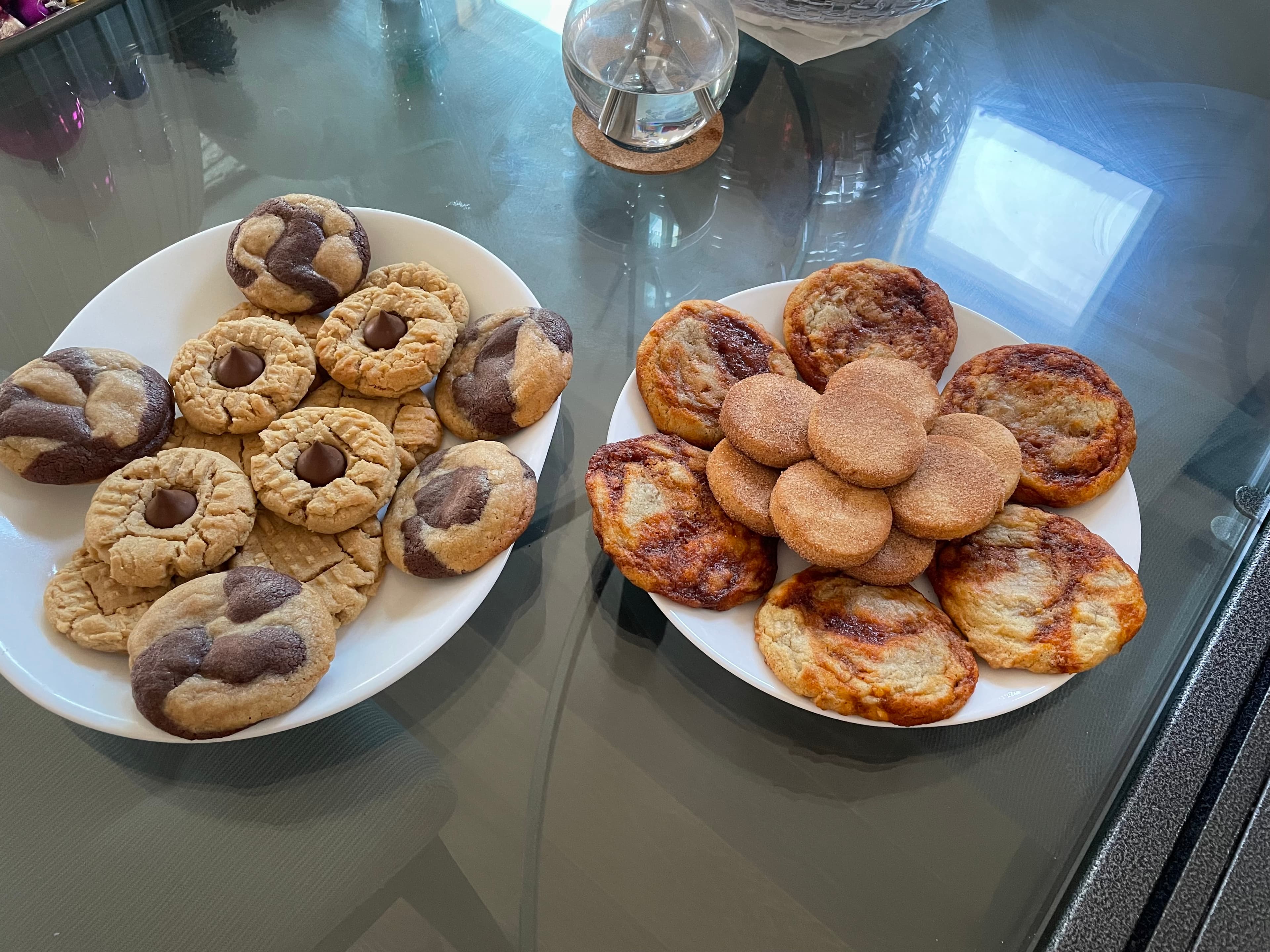 Four different kinds of cookies are piled on top of two white plates. The left plate has cookies with peanut butter and chocolate, and the right plate has cookies with spices like cinnamon and gochujang.